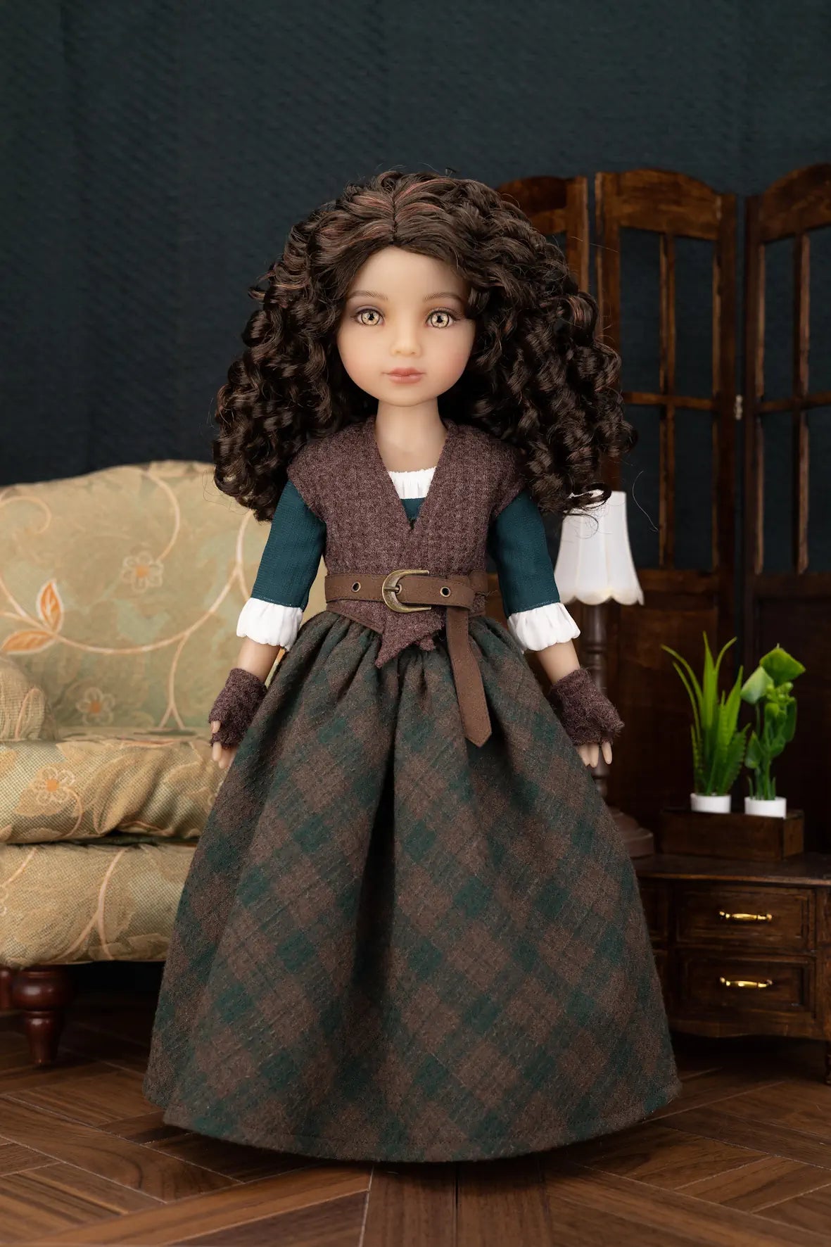 A doll with long curly brown hair wearing a detailed historical-style dress with a dark green and brown plaid skirt, a brown vest, and a dark teal top, posed in a miniature room setting.