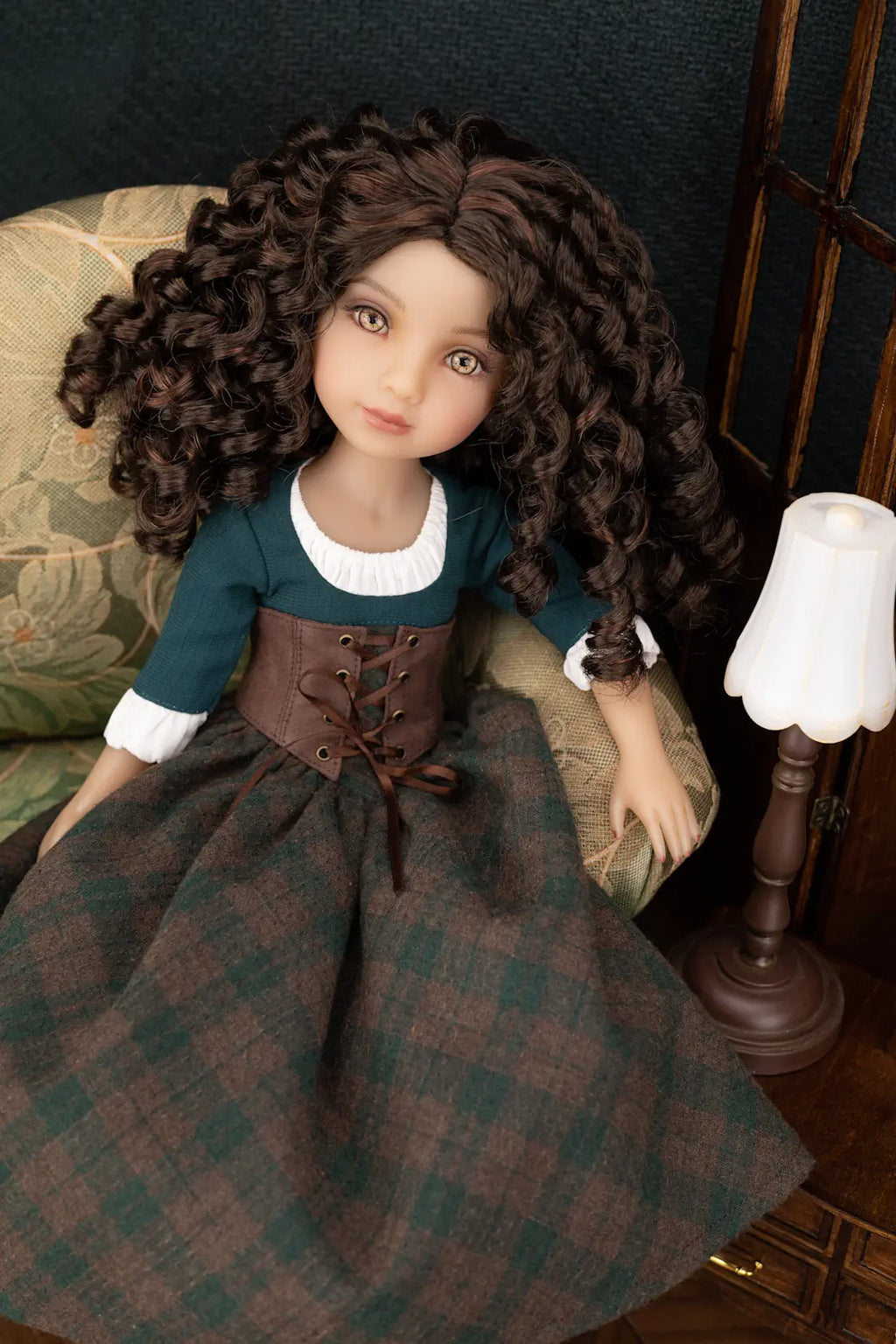 A doll with long curly brown hair sits on an upholstered chair next to a table lamp. The doll wears a dark teal dress with a white collar, a brown lace-up corset, and a brown plaid skirt.