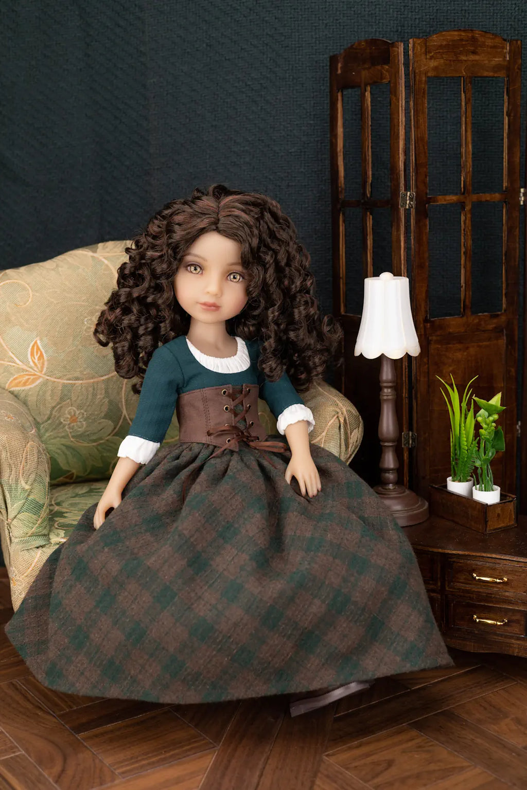 A doll with long curly brown hair sits on an upholstered armchair in a styled room scene. The doll wears a dark teal top with white cuffs, a brown corset-style waist, and a large dark green and brown plaid skirt.