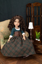 A doll with long curly brown hair sits on an upholstered armchair in a styled room scene. The doll wears a dark teal top with white cuffs, a brown corset-style waist, and a large dark green and brown plaid skirt.