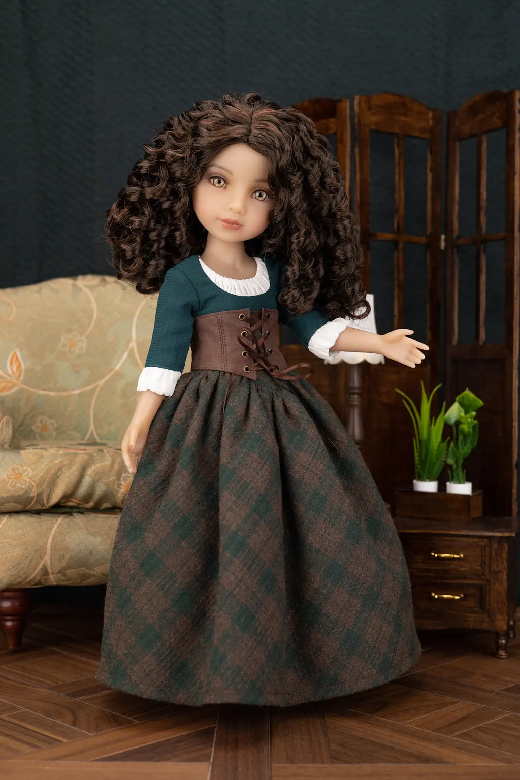 A doll with long curly brown hair wearing a dark teal and brown corset dress with a green plaid skirt, posed in a miniature room setting with a wooden screen, small table, and plant.