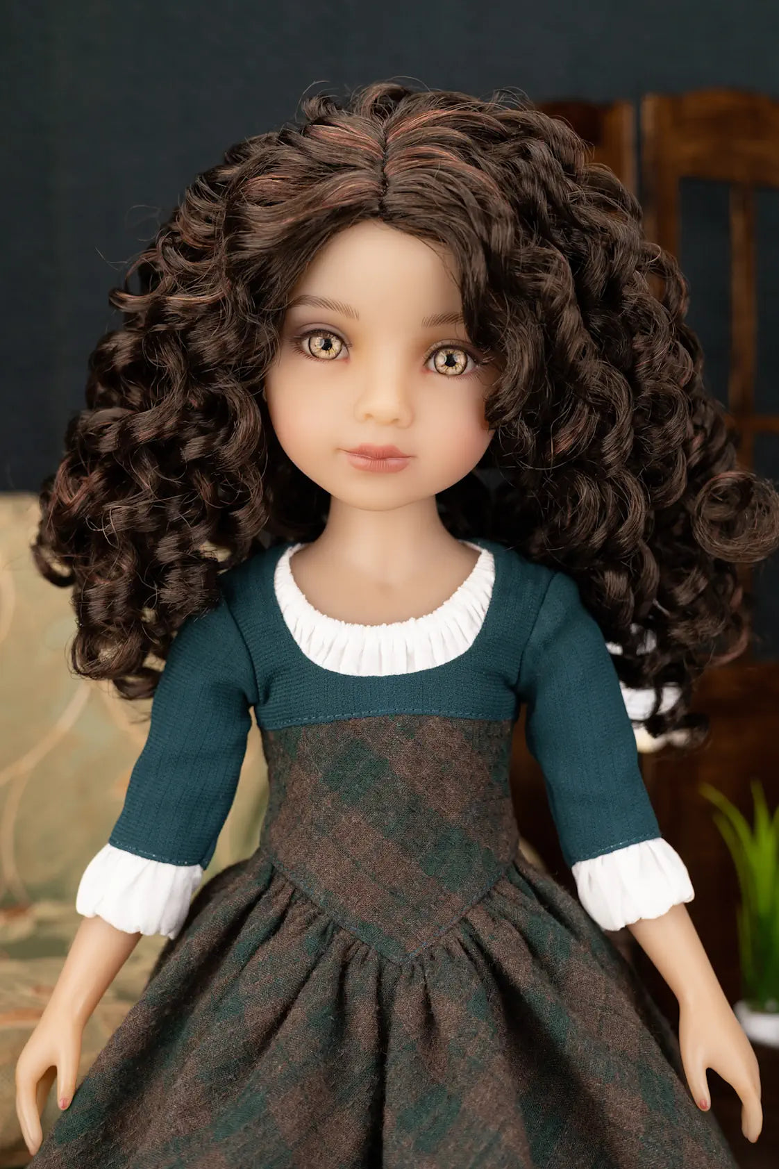 A doll with long, curly brown hair wearing a dark teal and brown dress with white ruffled trim, shown against a softly blurred indoor background.