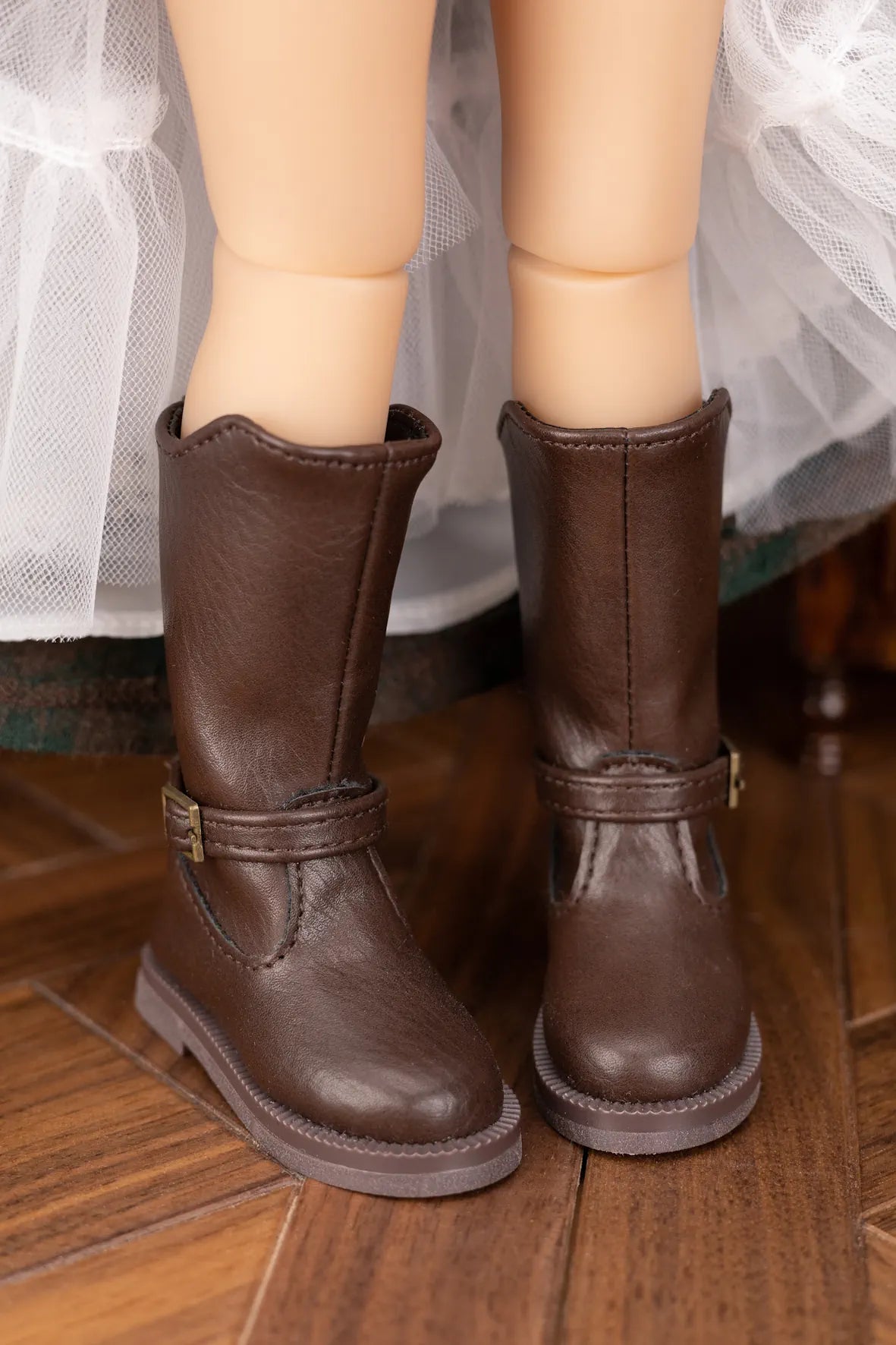 A pair of dark brown faux-leather boots with buckle straps, shown on a doll's legs standing on a wooden floor.