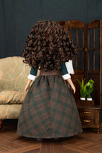 A doll viewed from behind wearing a long, voluminous dark brown curly wig, a dark green and brown plaid skirt, and a dark green top with white cuffs, standing in a miniature room setting.
