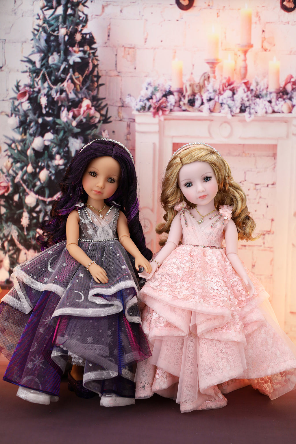 Two dolls in formal dresses standing in front of a decorated Christmas tree and fireplace.