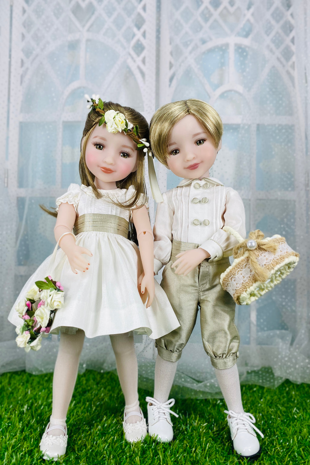 Two dolls, a girl in a white dress and a boy in formal attire, standing on grass with a decorative background.