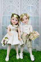 Two dolls, a girl in a white dress and a boy in formal attire, standing on grass with a decorative background.