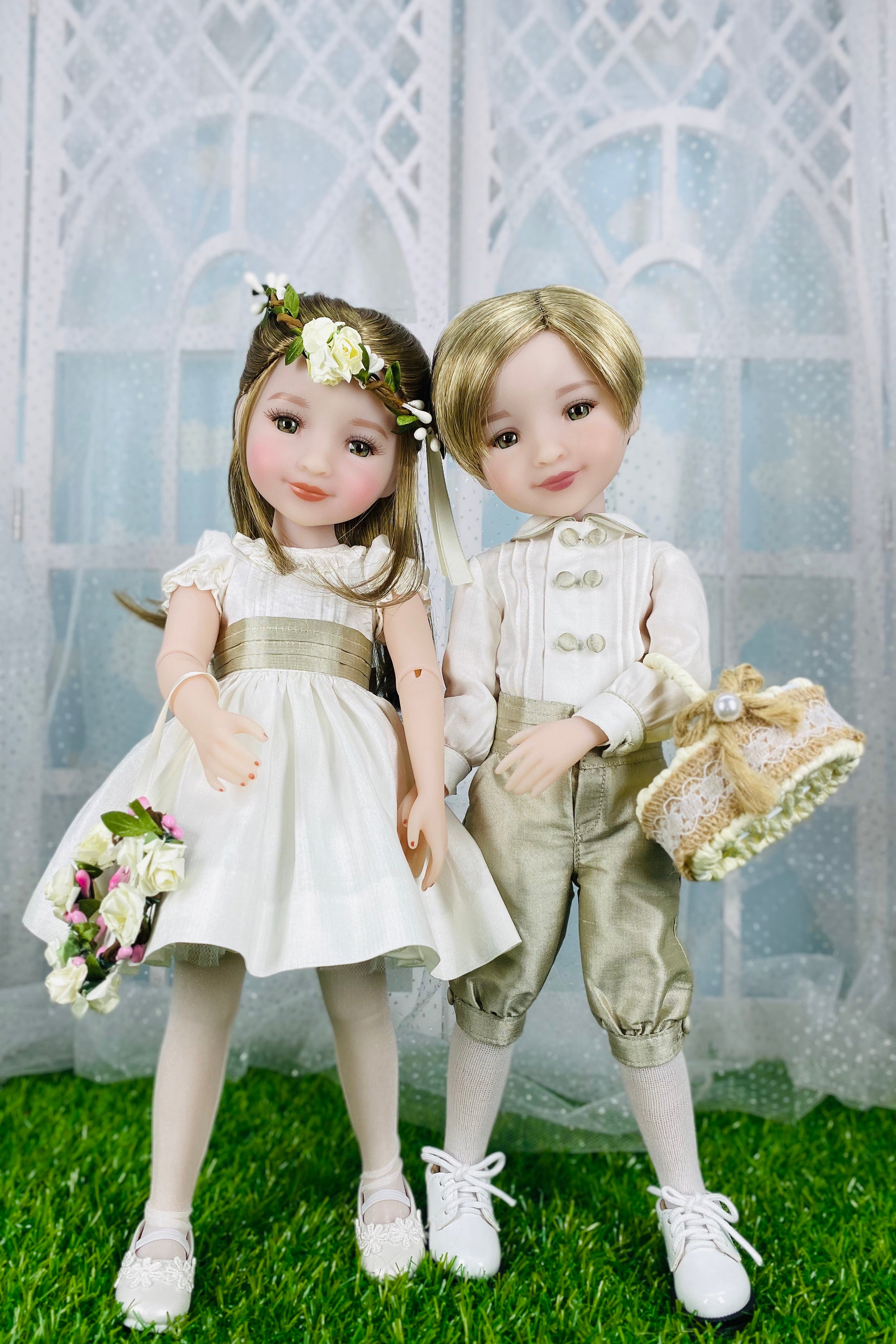 Two dolls, a girl in a white dress and a boy in formal attire, standing on grass with a decorative background.