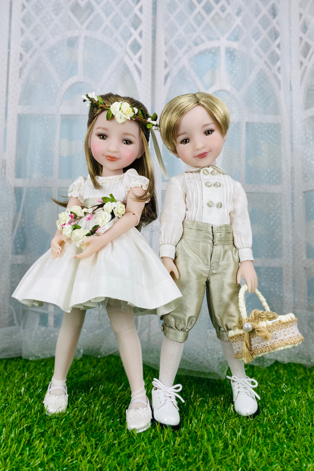 Two dolls, a girl and a boy, standing on grass with a decorative background.