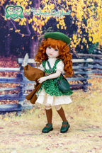 Doll with red hair and green outfit standing in a scenic outdoor setting with a wooden fence and trees.