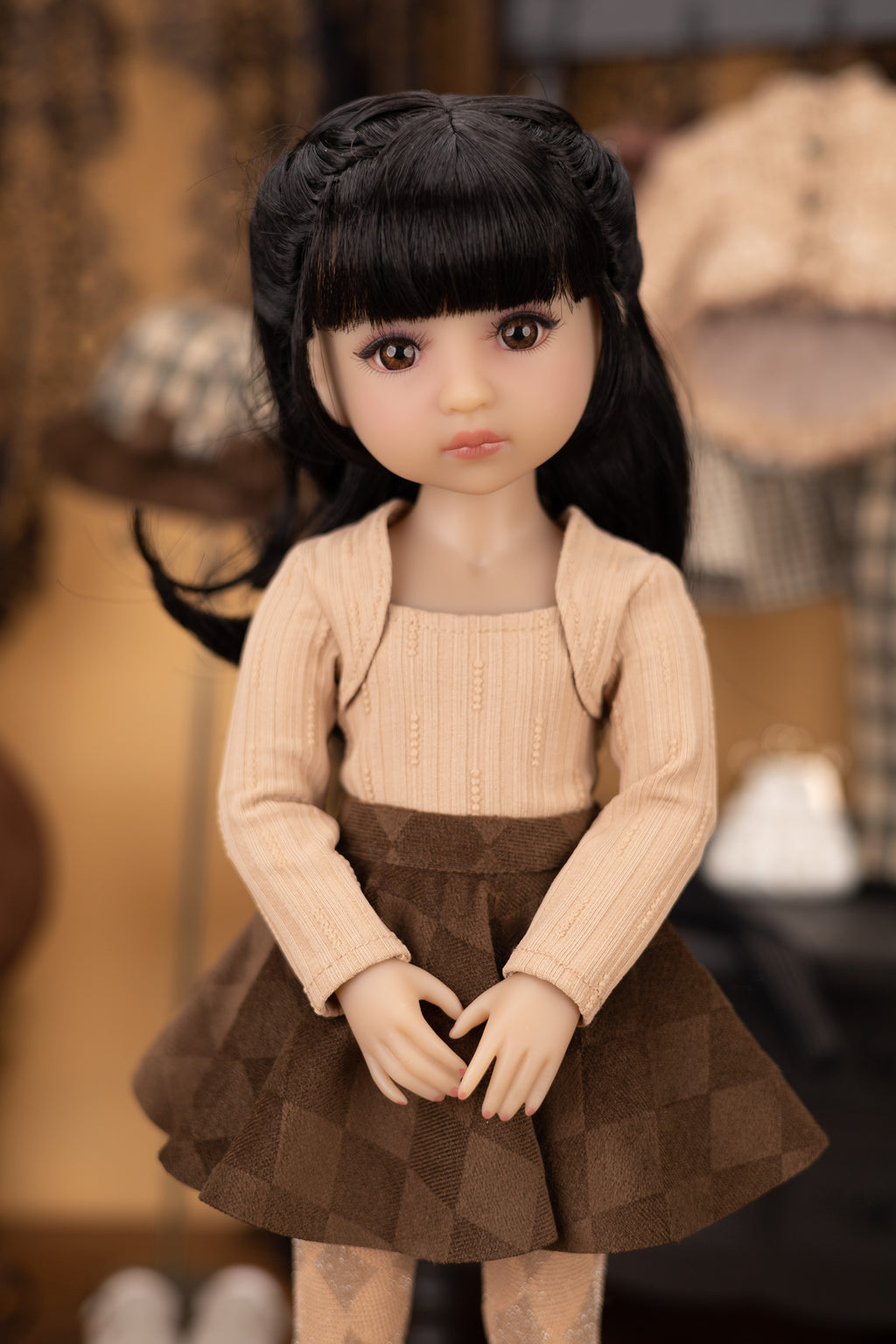 Doll with black hair and a beige sweater and brown skirt in a blurred indoor setting