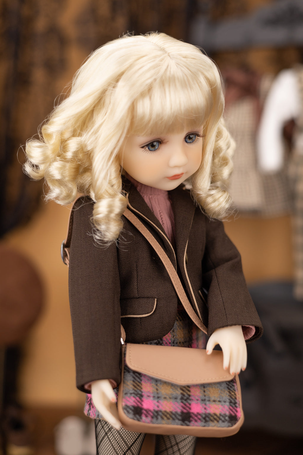 Doll with blonde hair wearing a brown coat holding a plaid handbag.