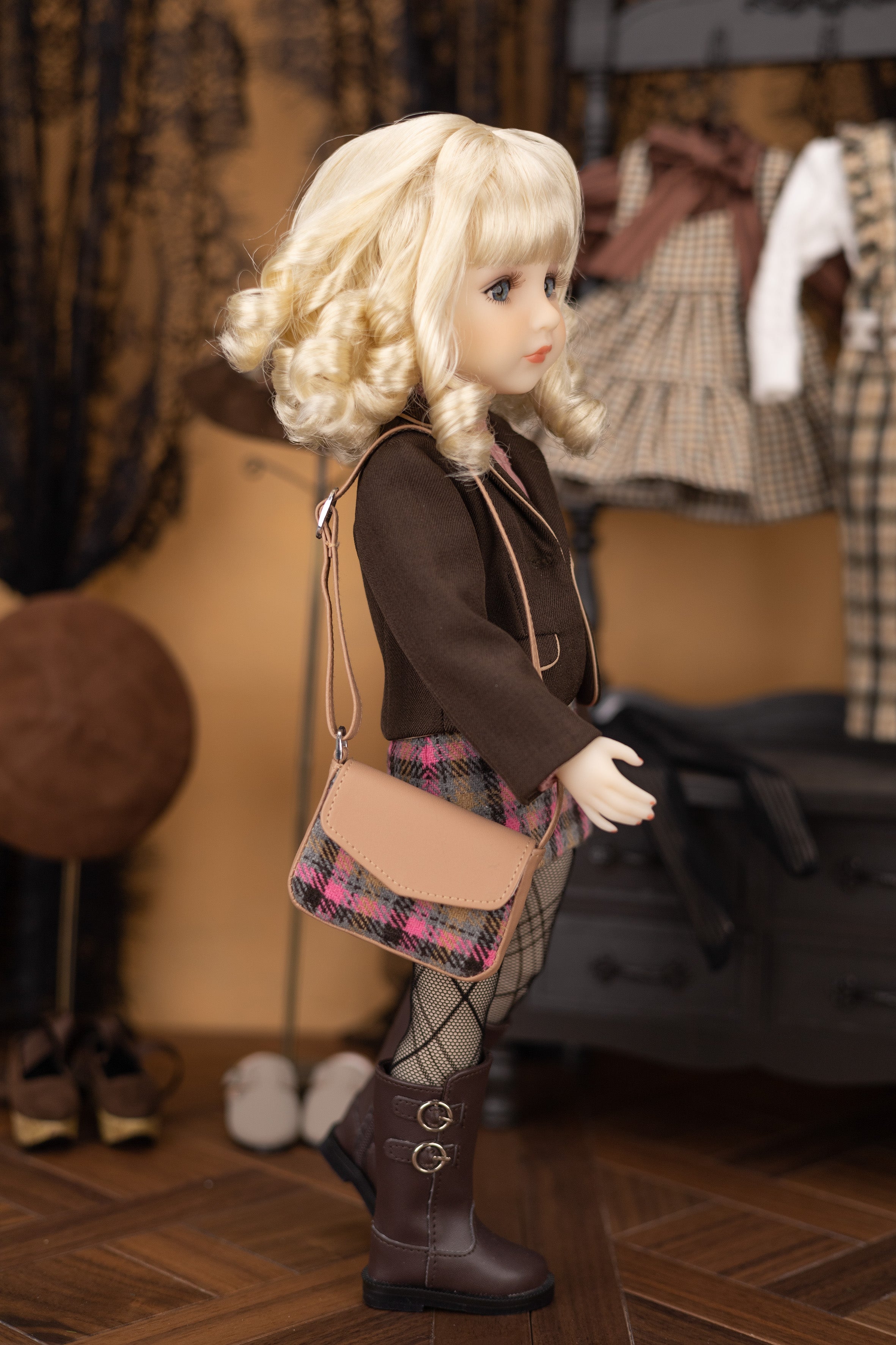 Doll with blonde hair, brown coat, plaid skirt, and brown boots holding a beige handbag in a room with clothing in the background.