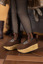 Brown shoes with cork soles worn by a person in striped tights on a wooden floor.