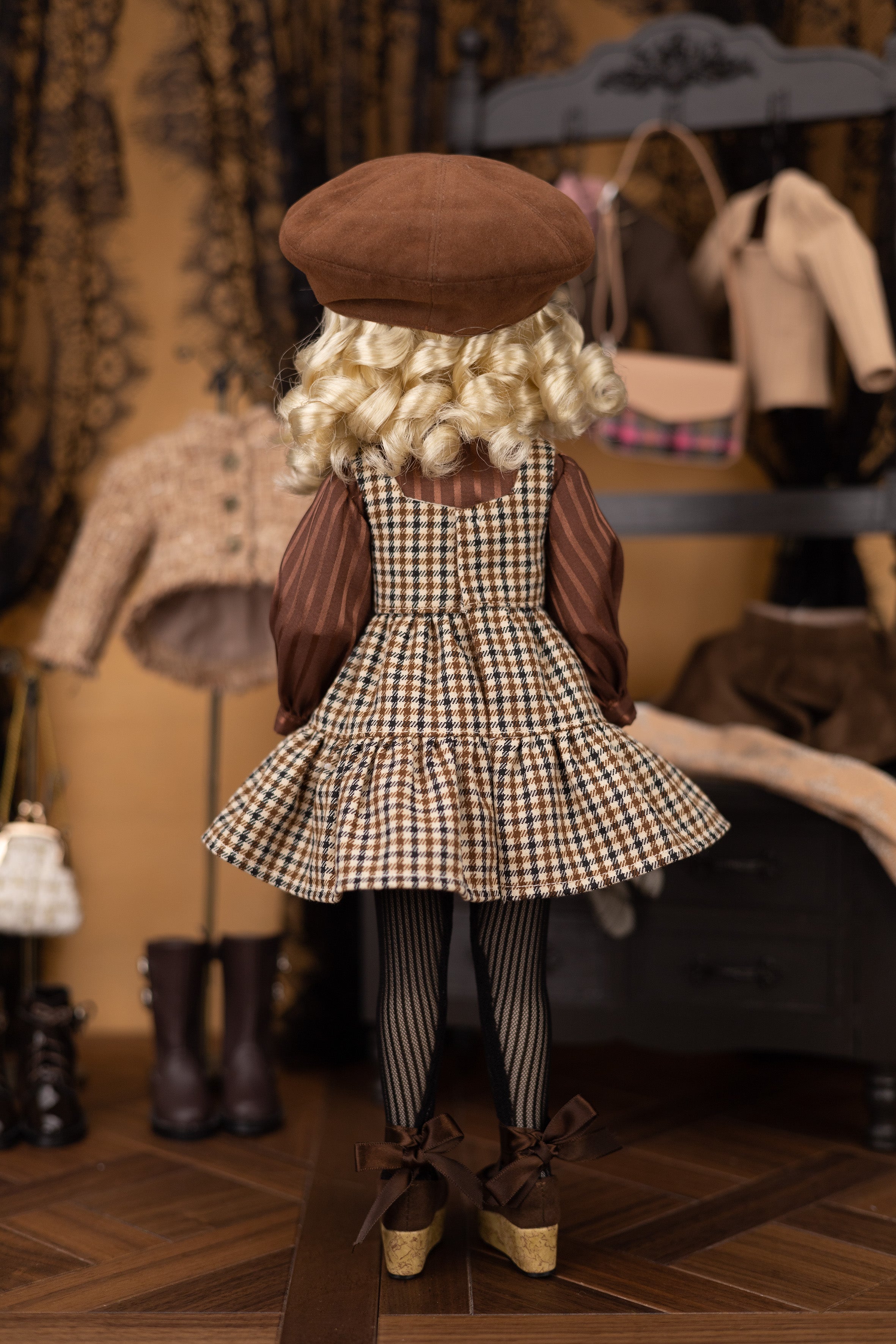 Doll in a checkered dress and brown hat standing in a room with clothing and furniture.