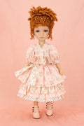 A doll with curly orange-brown hair wearing a pink floral tiered dress with white lace trim and pink lace-up shoes, standing against a solid pink background.