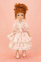 A doll with curly orange-brown hair wearing a pink floral tiered dress with white lace trim and pink lace-up shoes, standing against a solid pink background.