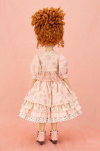 A doll viewed from behind, wearing a light pink floral tiered dress with white polka-dot trim and pink shoes, standing against a solid pink backdrop.