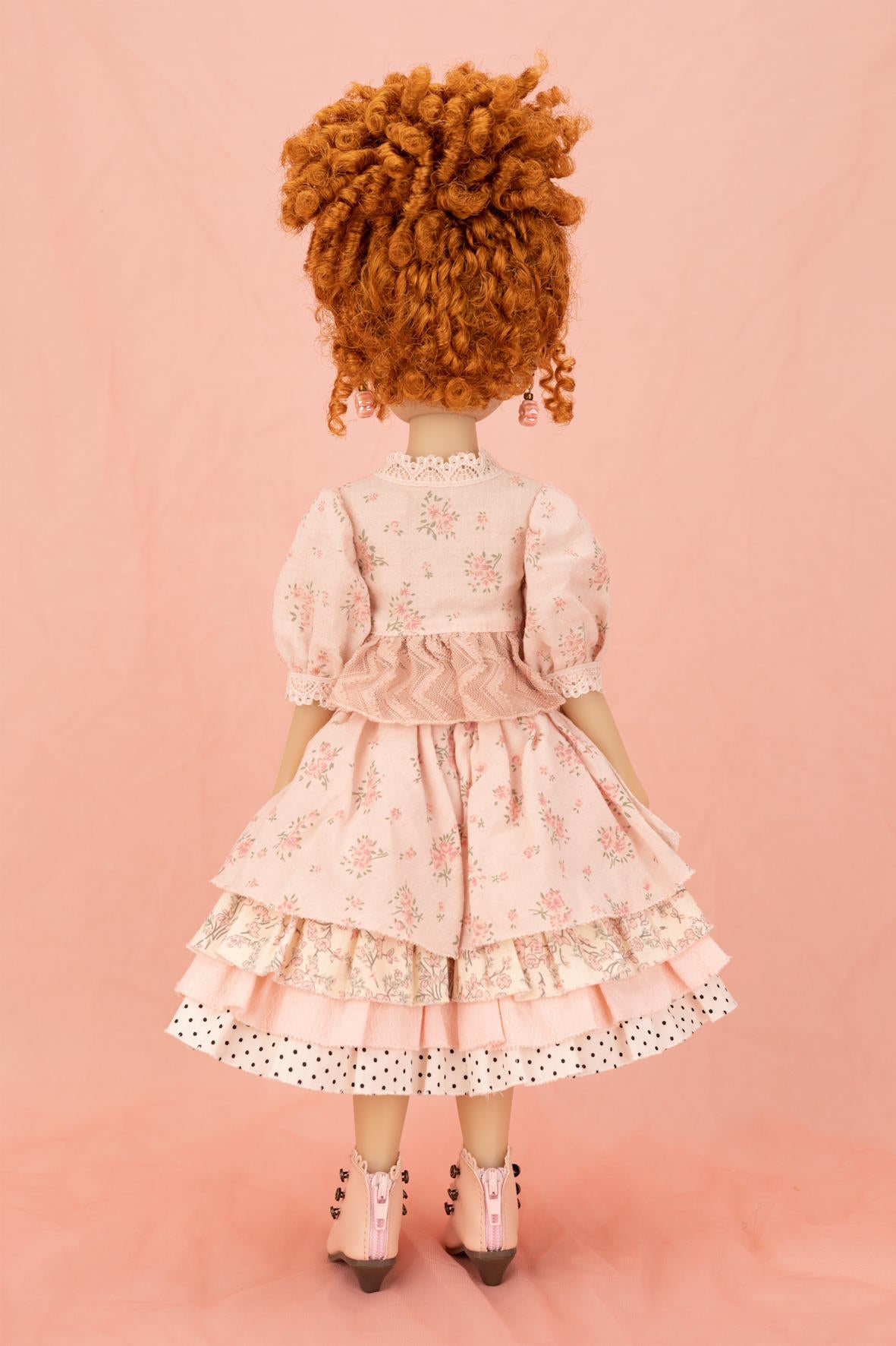 A doll viewed from behind, wearing a light pink floral tiered dress with white polka-dot trim and pink shoes, standing against a solid pink backdrop.