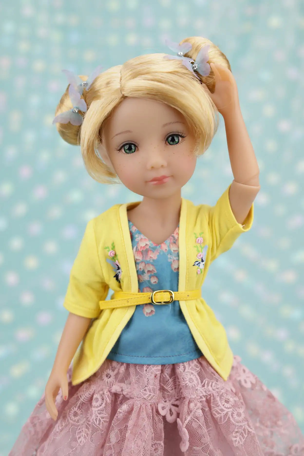 Juniper with blonde hair styled in pigtails, wearing a yellow cardigan, blue top, and pink skirt against a light blue polka dot background.