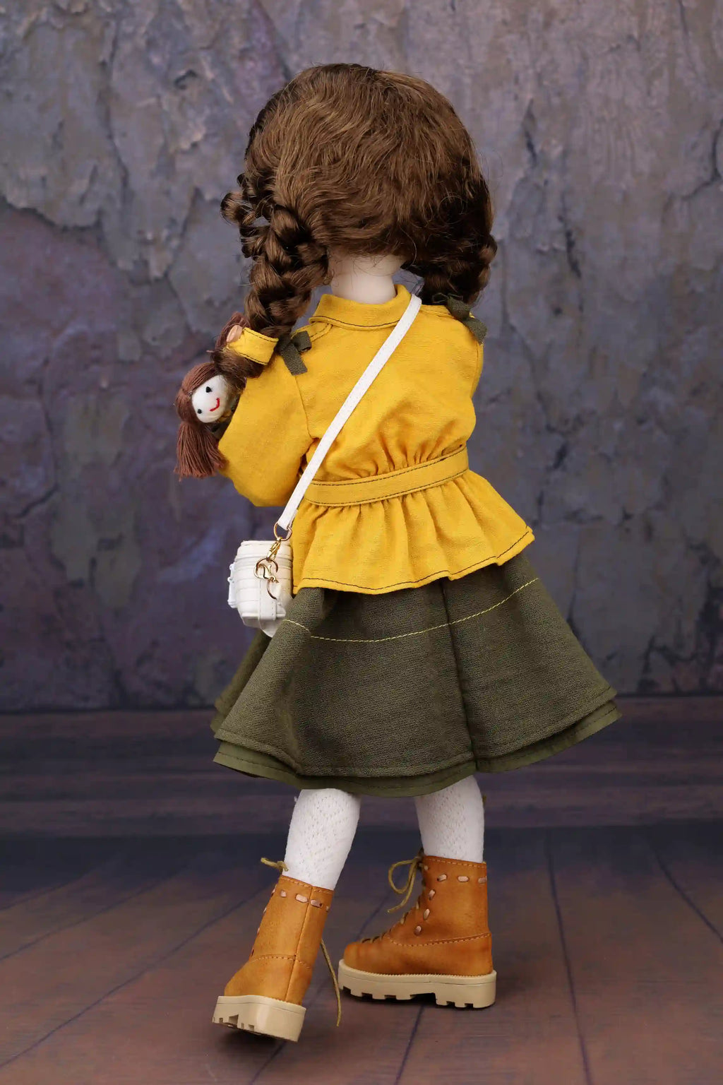 Doll wearing a yellow top and green skirt with brown boots, standing against a textured wall.