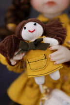 Small doll with brown hair and a yellow dress holding a green bow.