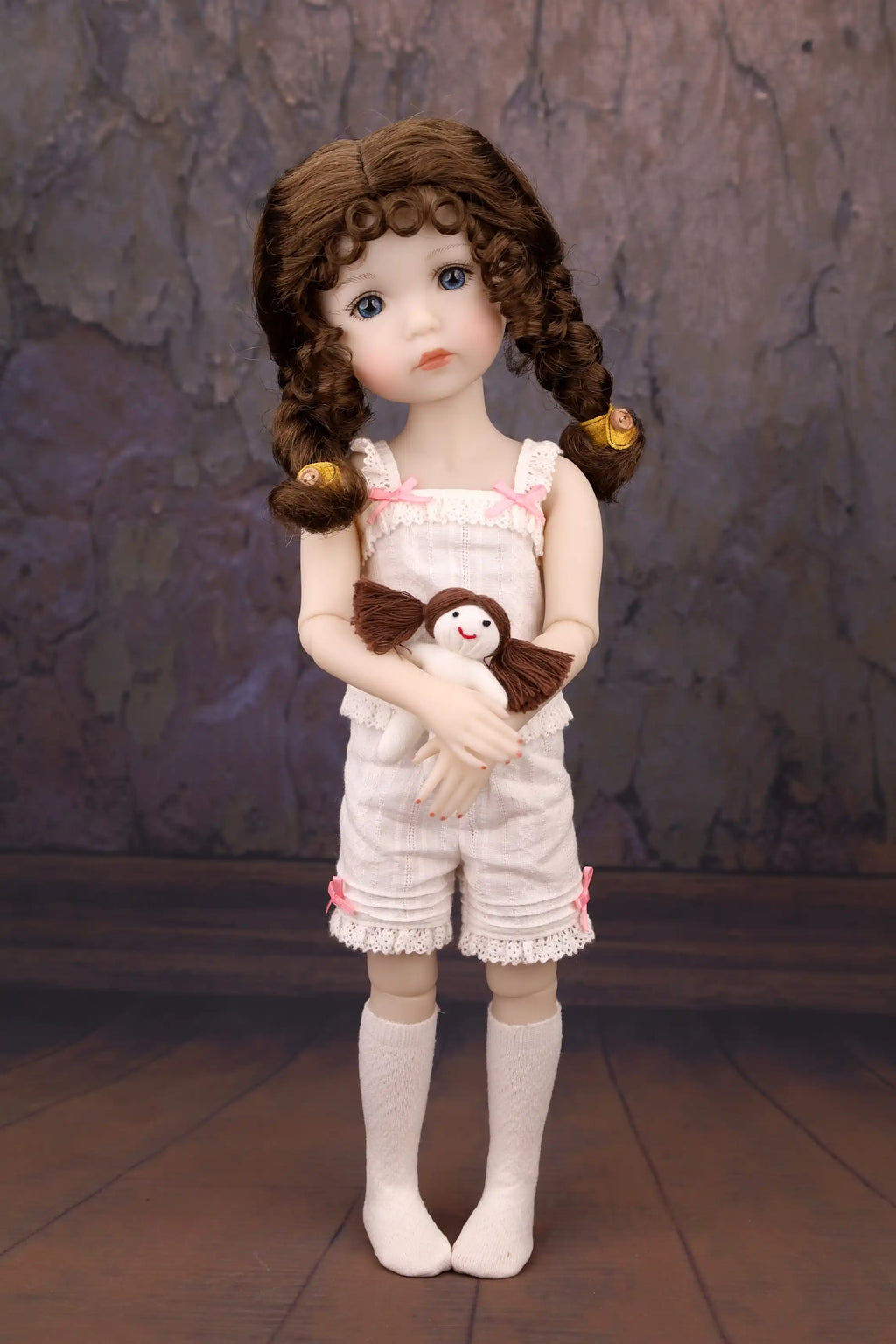 Doll with curly hair wearing a pink outfit holding a teddy bear against a textured wall.