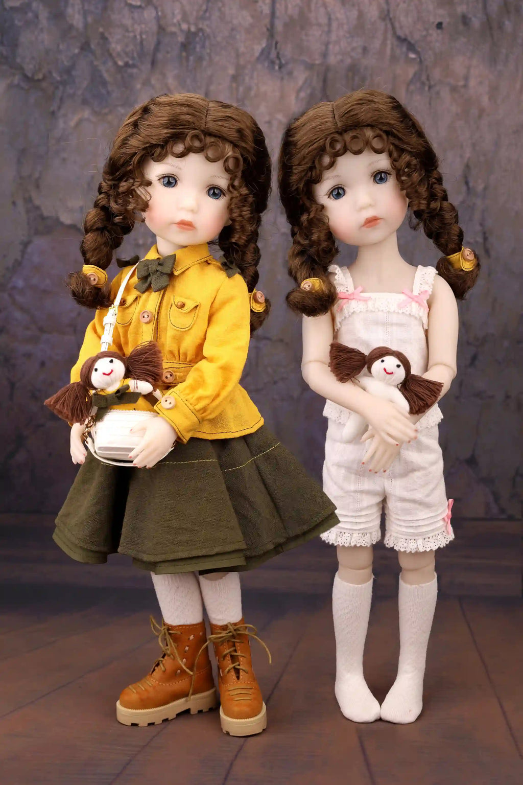 Two dolls with braided hair, one in a yellow outfit and the other in a white outfit, holding small stuffed animals against a textured wall.