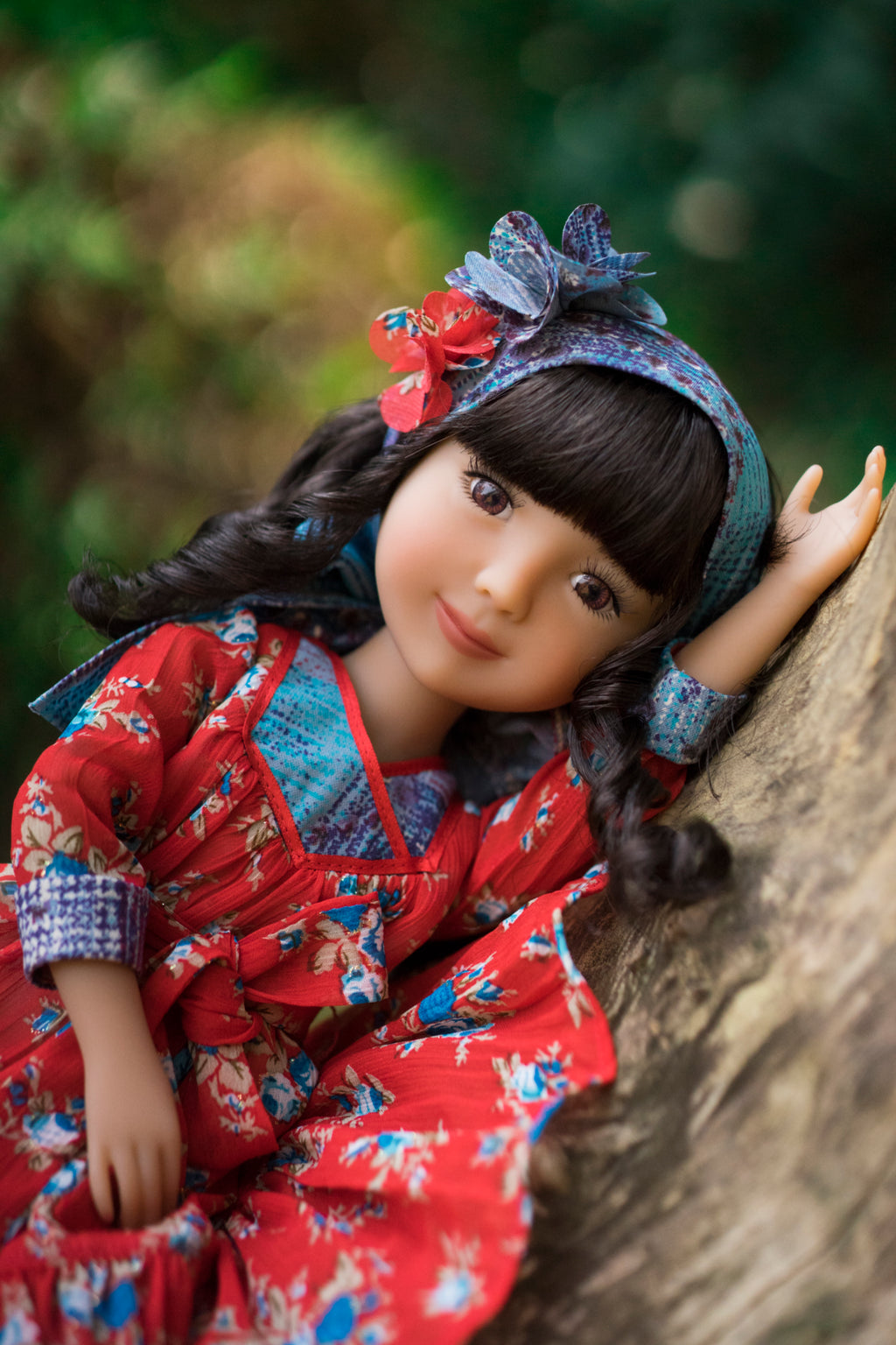 Doll in a red outfit with floral patterns sitting against a tree trunk outdoors.