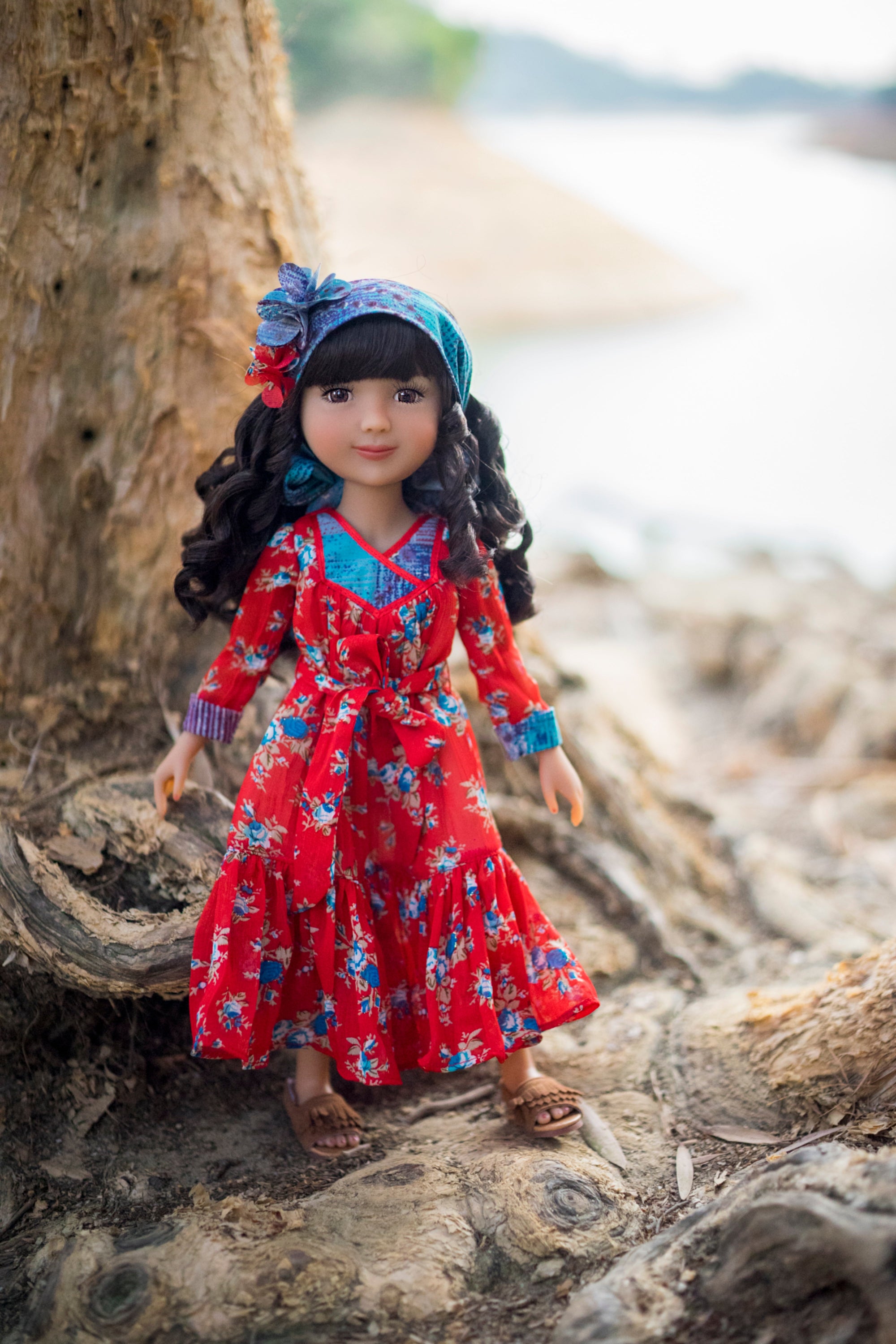 Doll in a red floral dress and blue headscarf standing on rocks near a tree