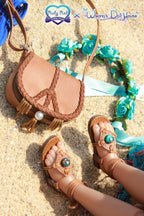 Pair of brown sandals with decorative elements on a sandy surface, featuring 'Ruby Red' and 'Whimsy's Doll House' logos.