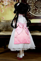 Doll with braided hair wearing a white dress with a pink bow in a decorative setting.