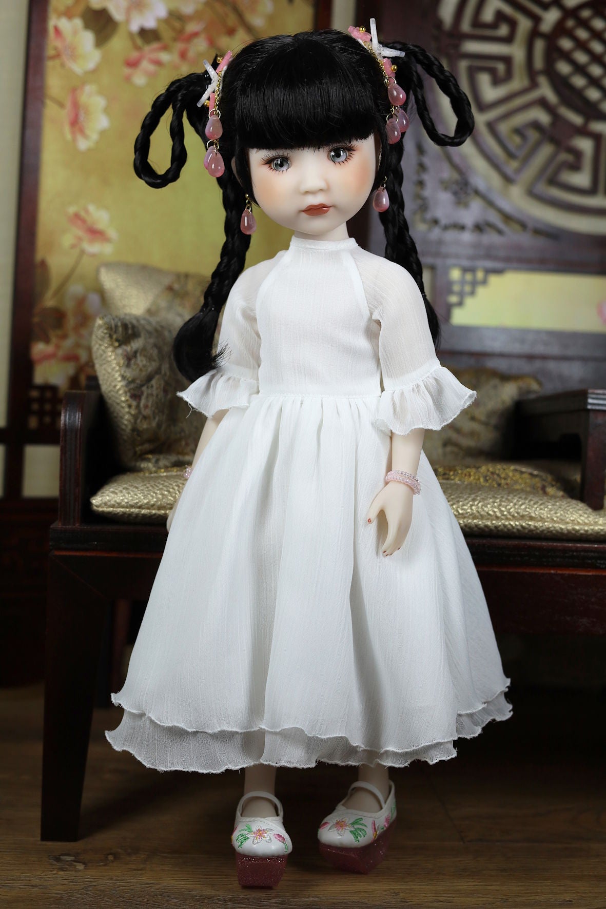 Doll in a white dress with black hair styled in pigtails, sitting on a wooden chair.