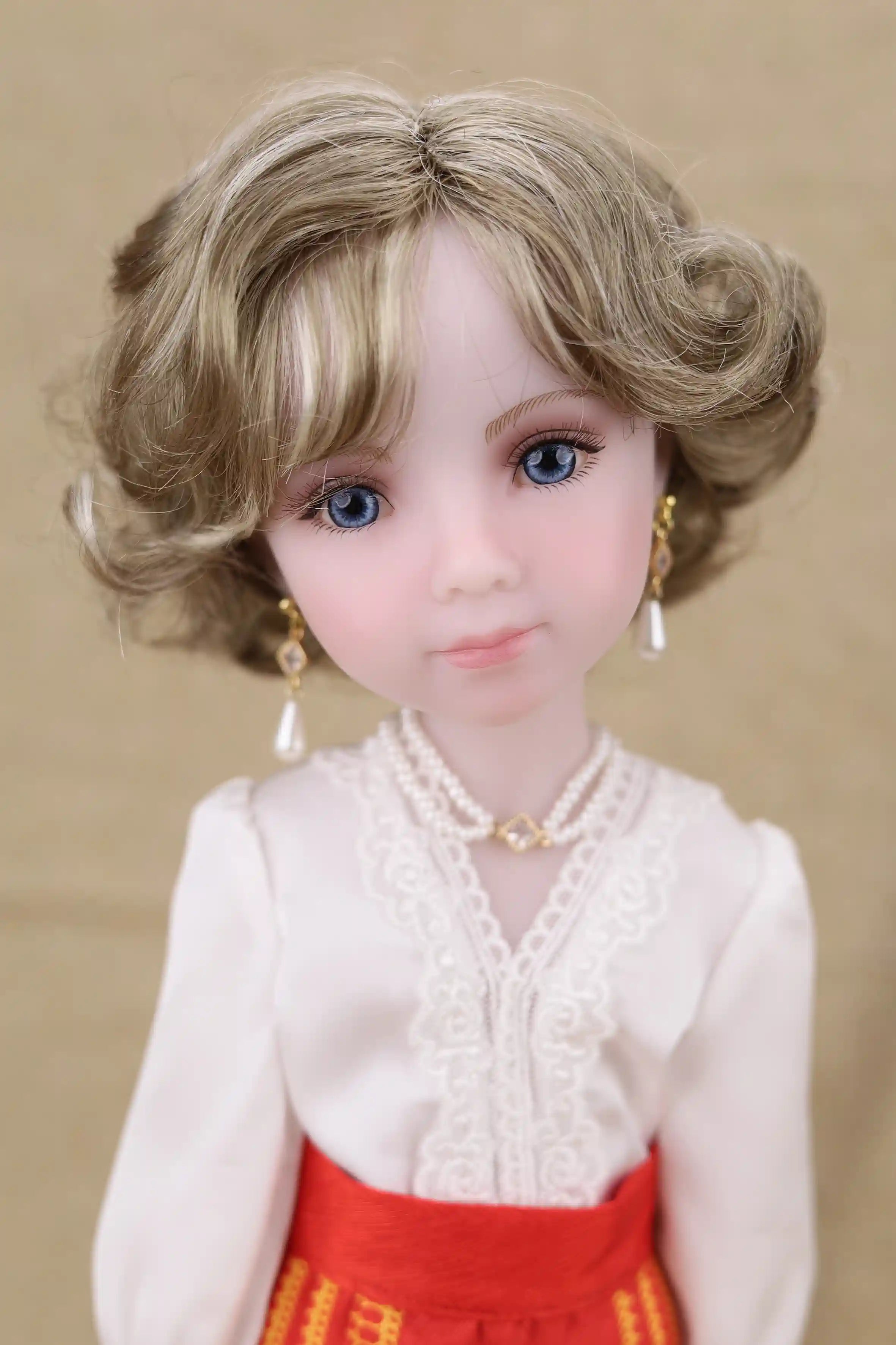 Doll with blonde hair and blue eyes wearing a white blouse and red skirt against a beige background