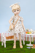 Doll in a floral dress standing in a garden setting with a bench and table.