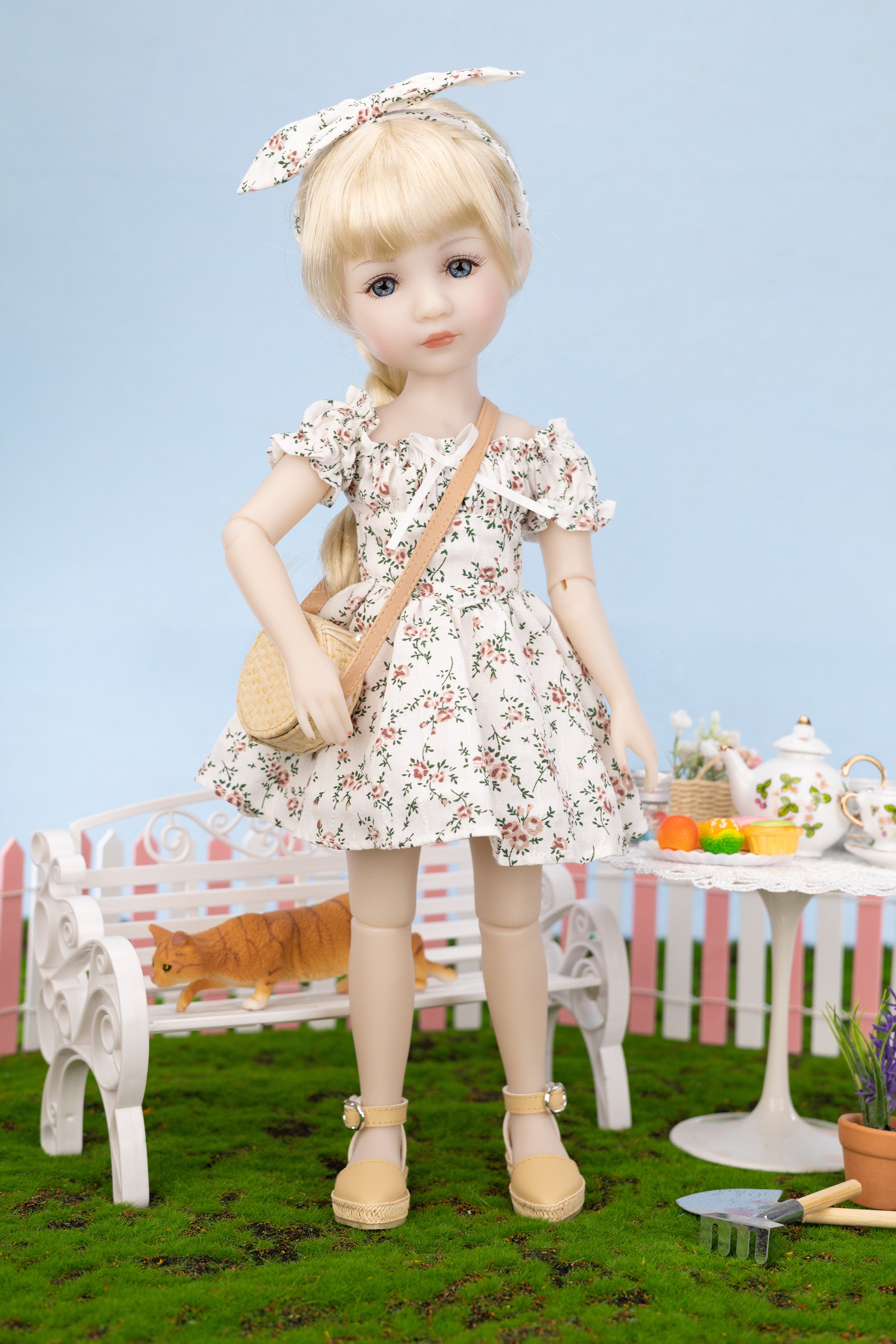 Doll in a floral dress standing in a garden setting with a bench and table.