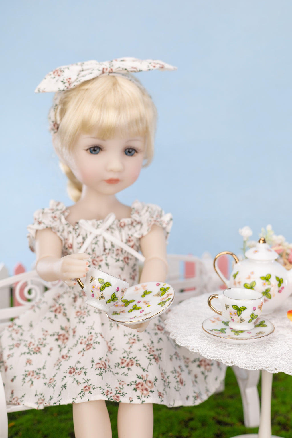 Doll in a floral dress holding a teacup with a tea set on a table against a blue background