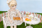 Doll in a floral dress with a cat, sitting at a small table with tea set and fruits in a garden setting.