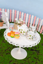 Miniature tea set and pastries on a white lace tablecloth with a picket fence and flowers in the background.