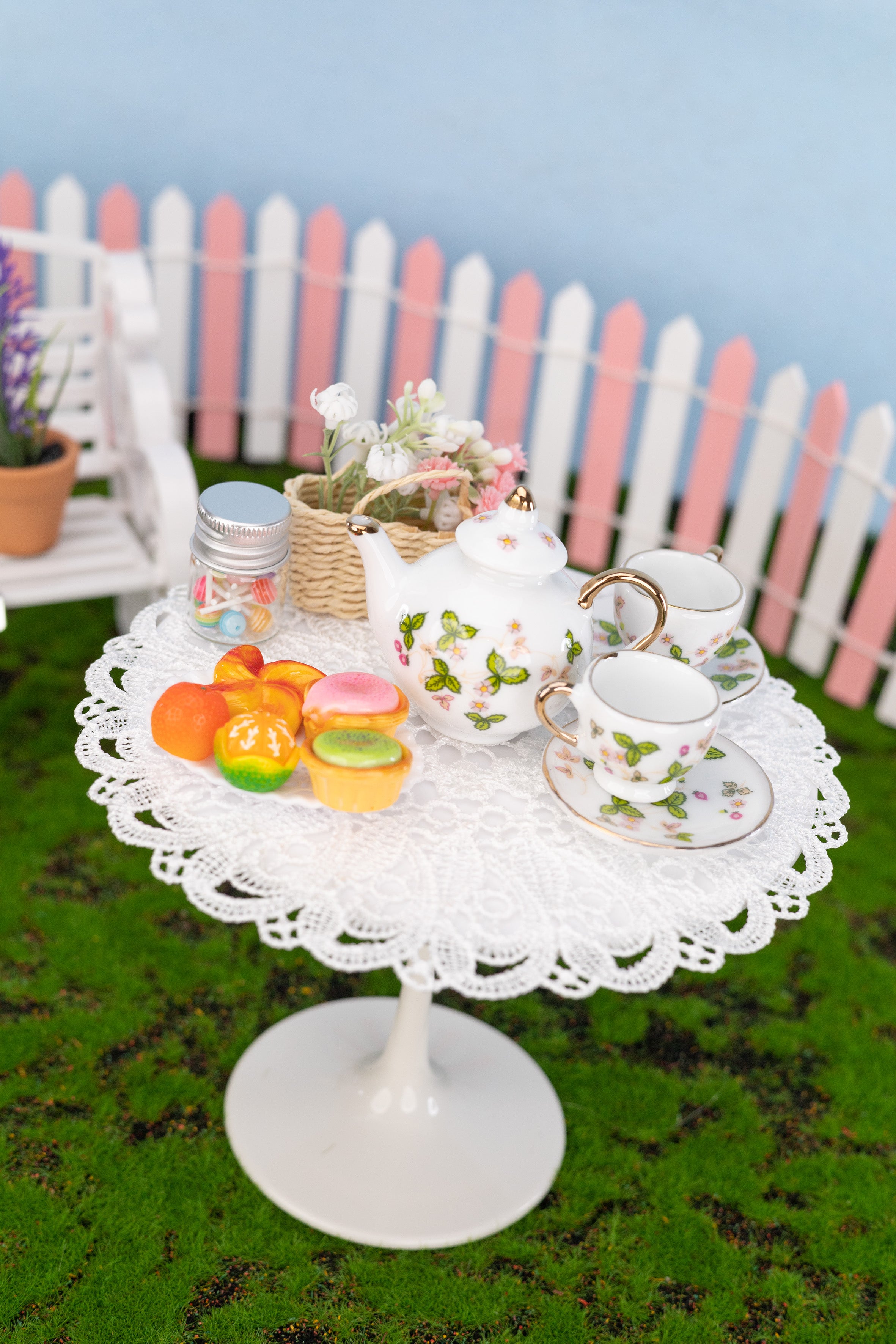 Miniature tea set and pastries on a white lace tablecloth with a picket fence and flowers in the background.