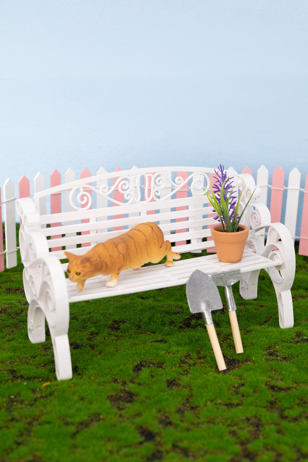 White bench with a cat and potted plant on a grassy area with a colorful fence in the background.