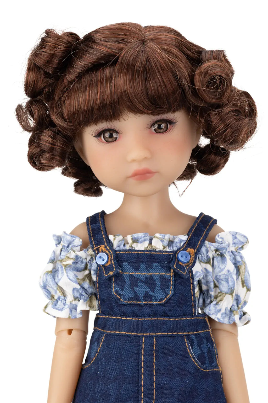 Doll with brown hair styled in buns, wearing a denim overalls and floral blouse on a white background