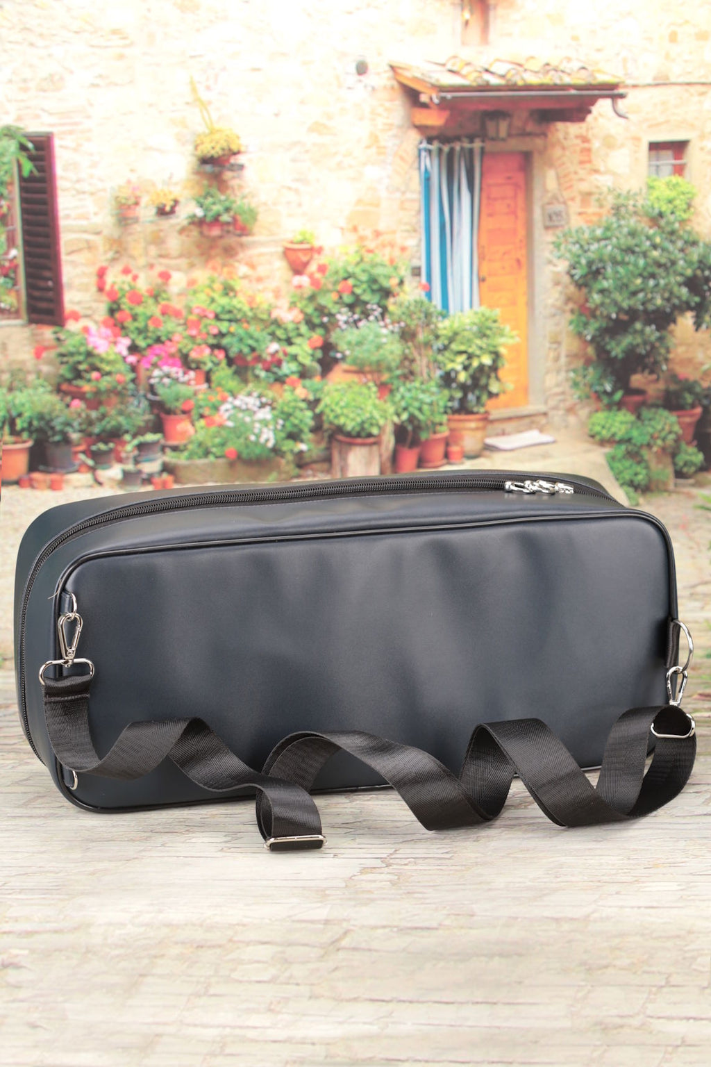 Black doll carry bag with straps on a wooden surface in front of a stone building with plants.