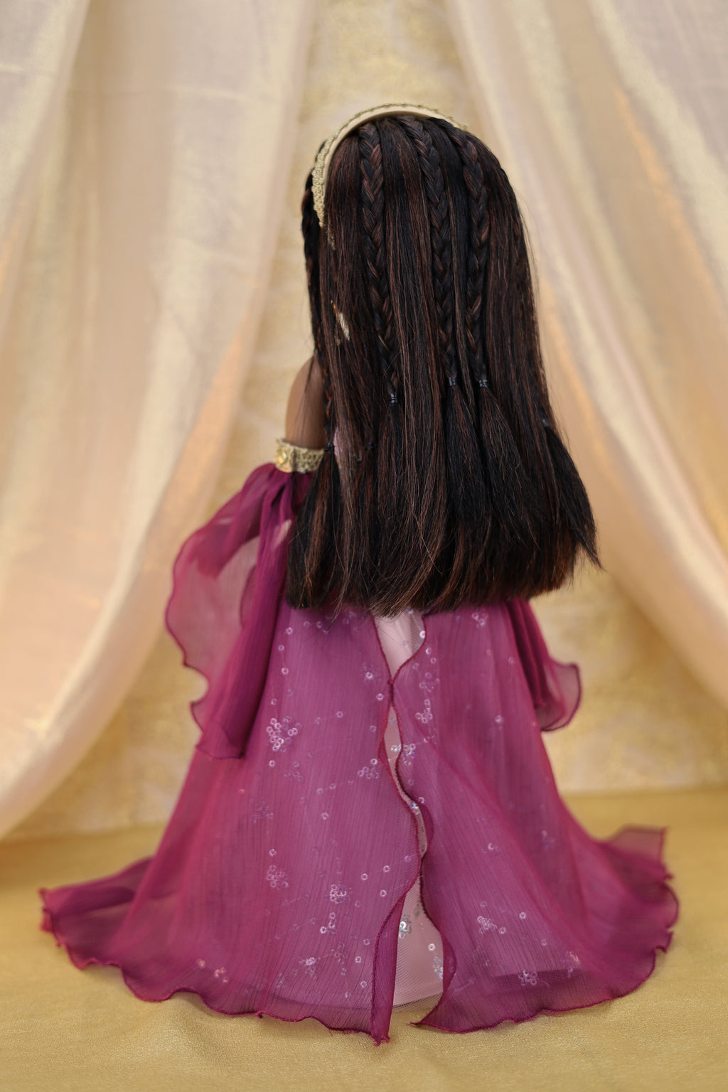 Doll with long braided hair wearing a purple outfit against a beige curtain background