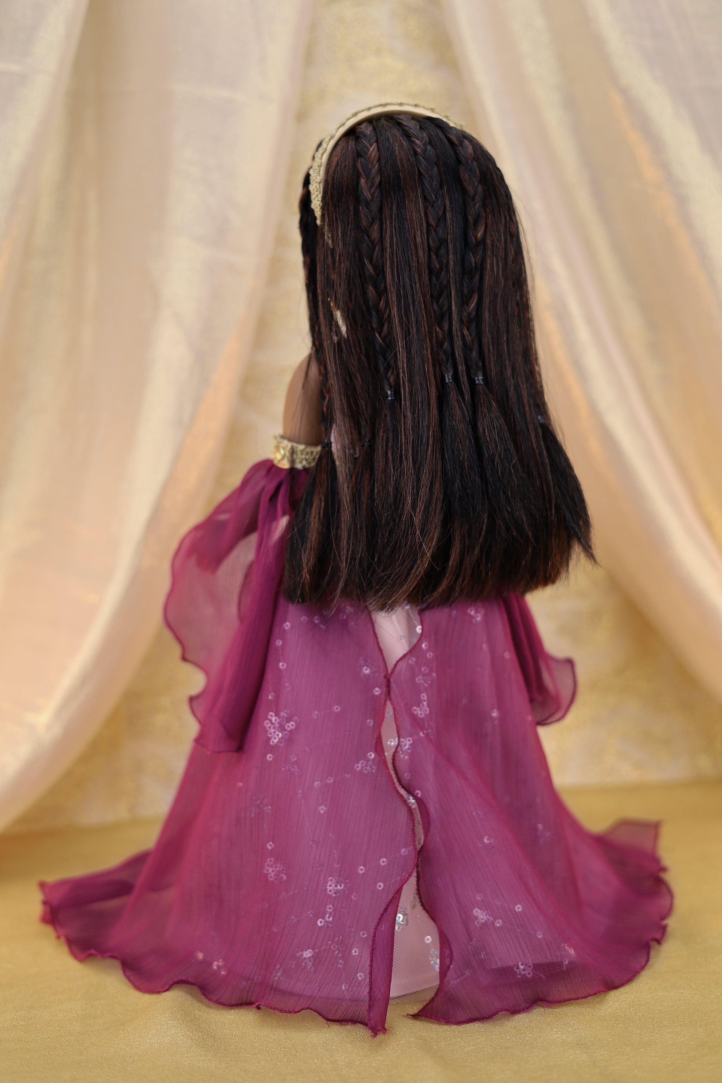 Doll with long braided hair wearing a purple outfit against a beige curtain background