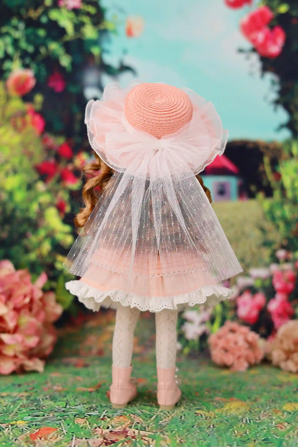 Doll wearing a large pink hat and dress in a garden setting with flowers and greenery.