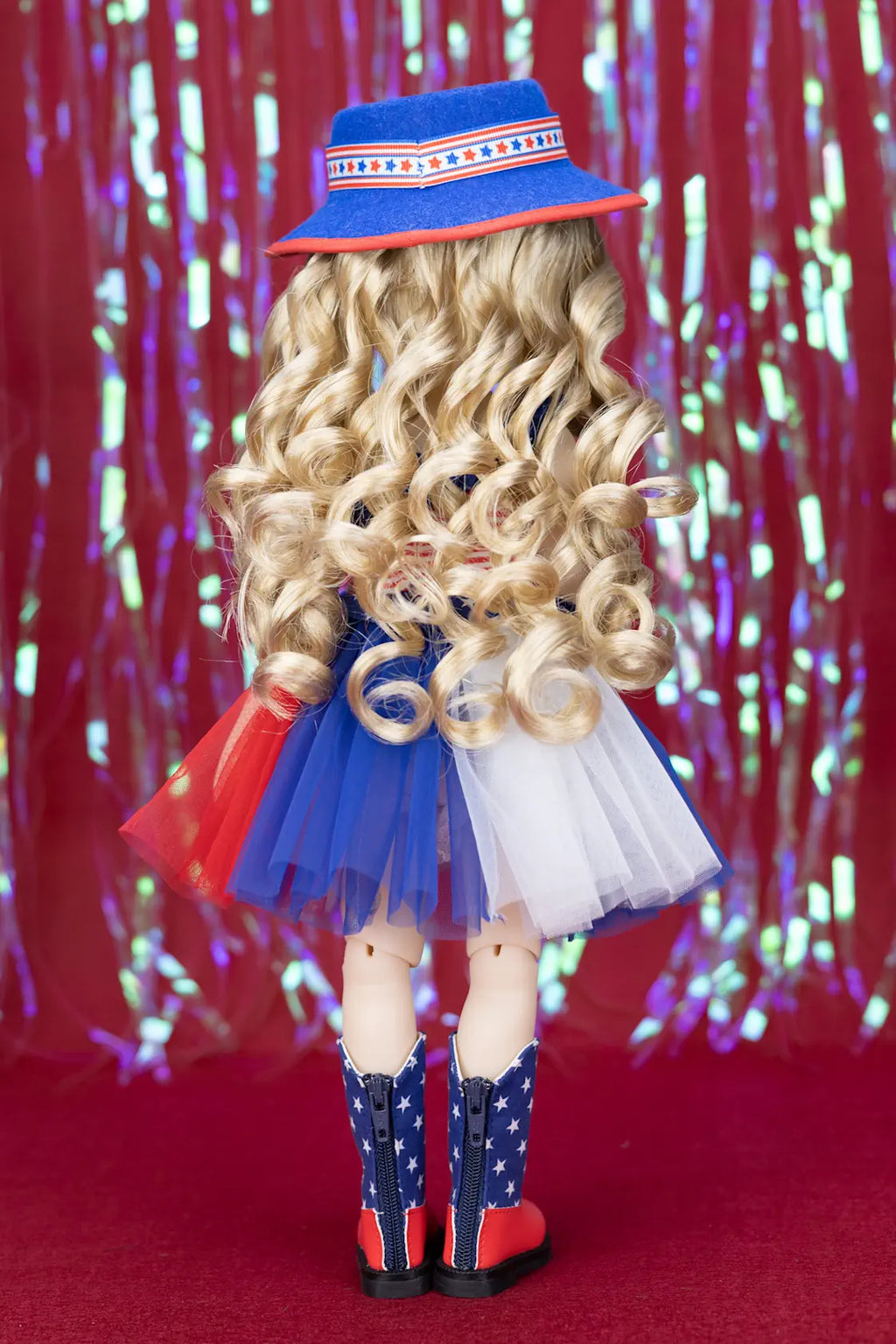 Doll with curly blonde hair wearing a blue hat, red skirt, white top, and star-patterned boots against a colorful curtain background.