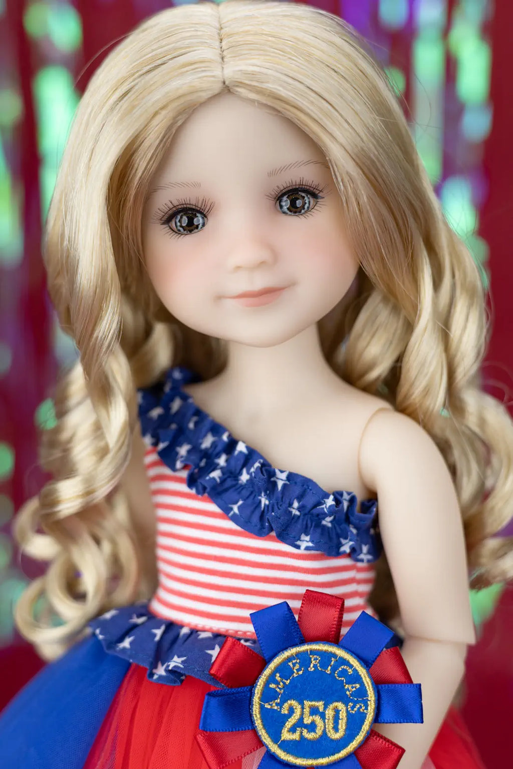 Doll wearing a patriotic outfit with a 'merica 250' ribbon against a colorful background