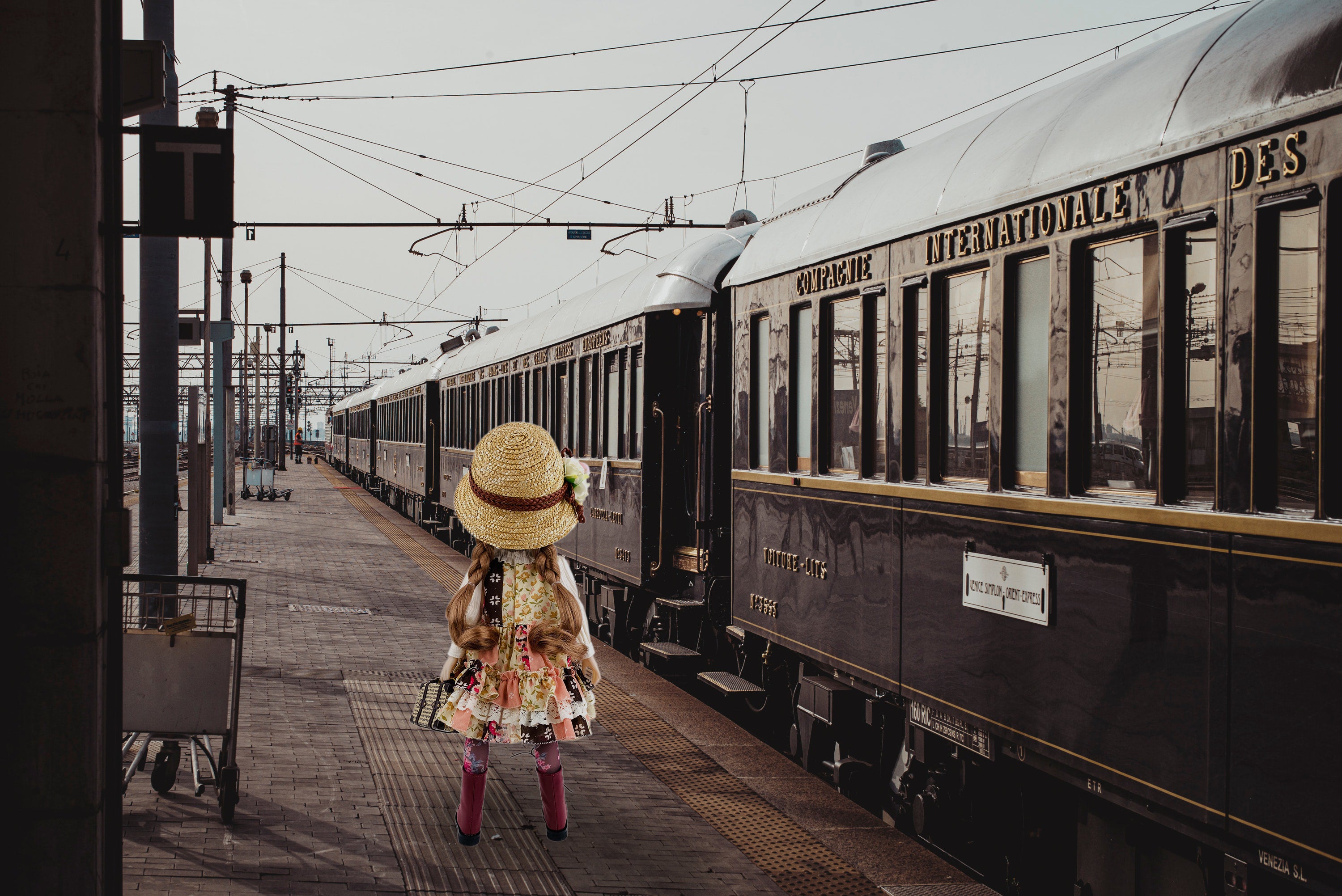 Doll in a vintage outfit standing on a train platform with an old-fashioned train.