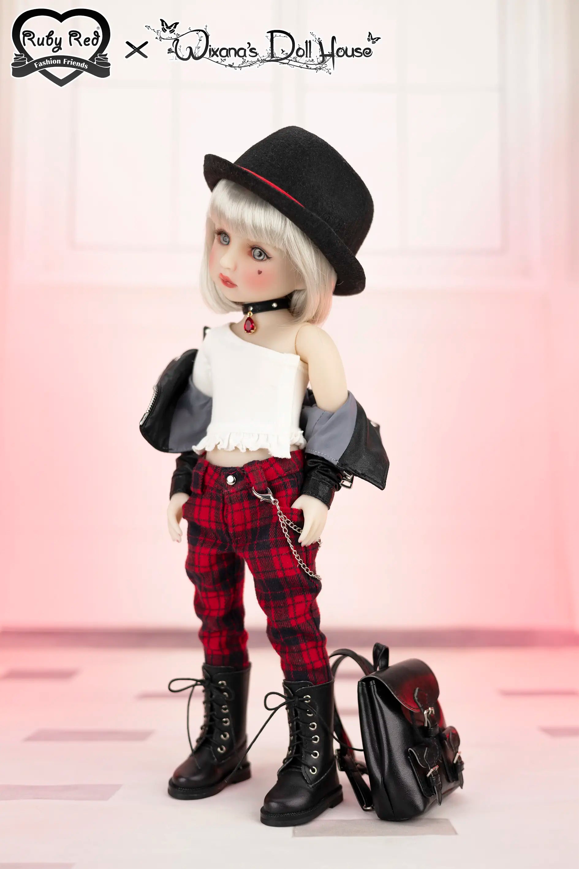 Doll with black hat, white top, red plaid pants, and black boots standing next to a backpack on a pink background.